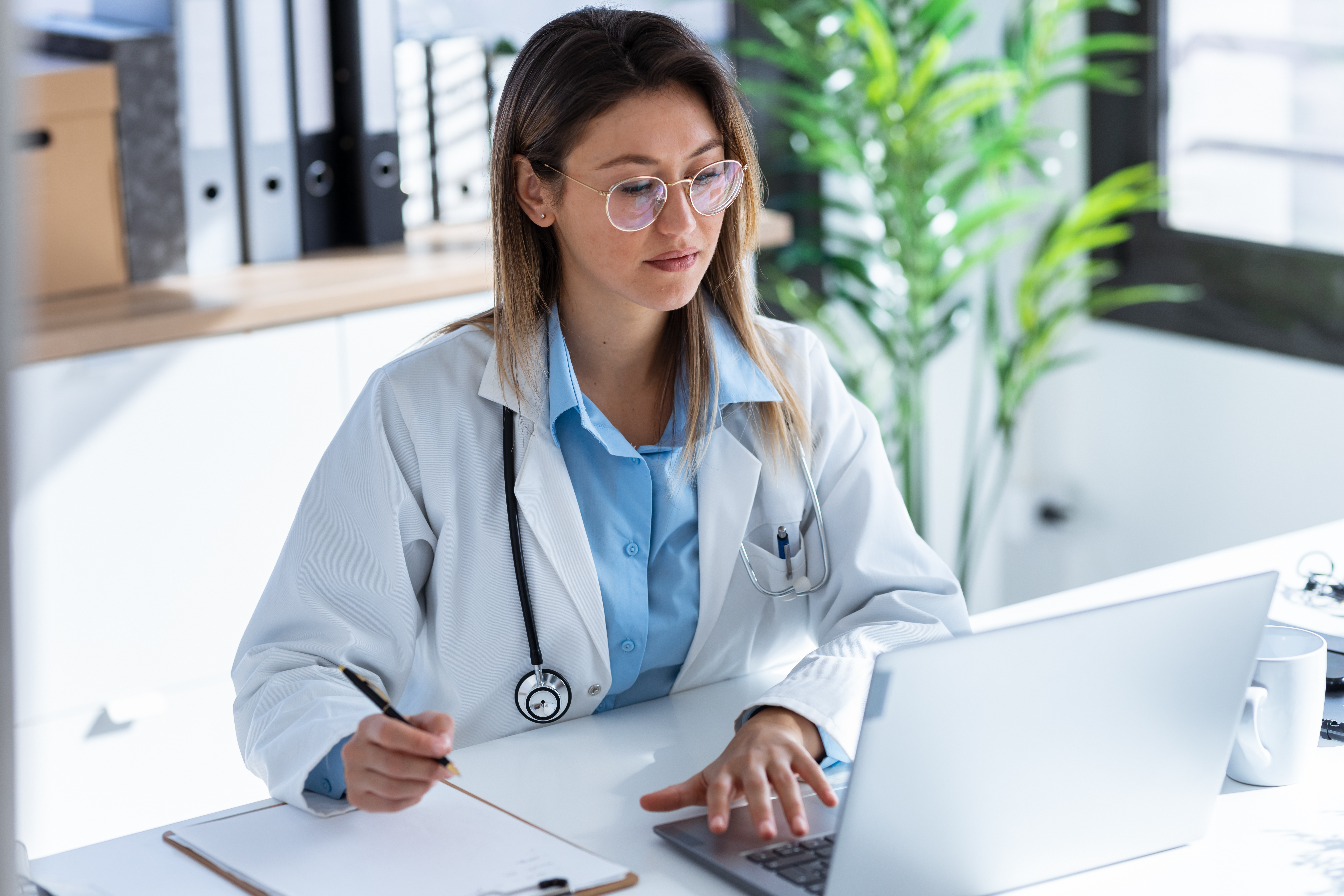 female-latin-american-adult-doctor-consultation-room-searching-on-laptop-while-writing-web